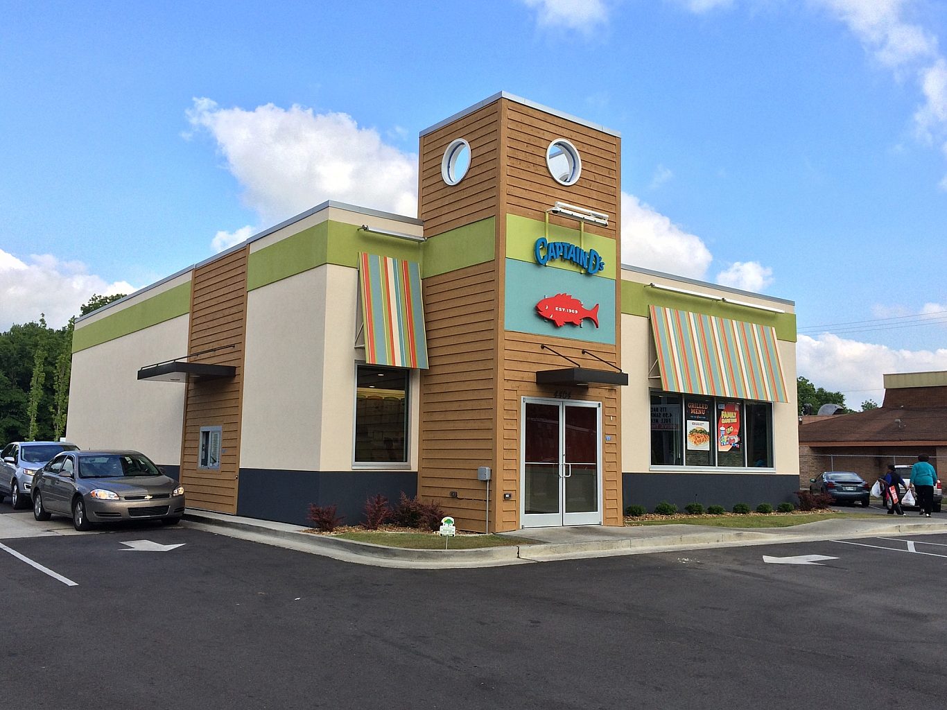 The exterior of a Captain D's restaurant. A red fish is above the entrance, and striped awnings are over the windows. A car is in the drive through.