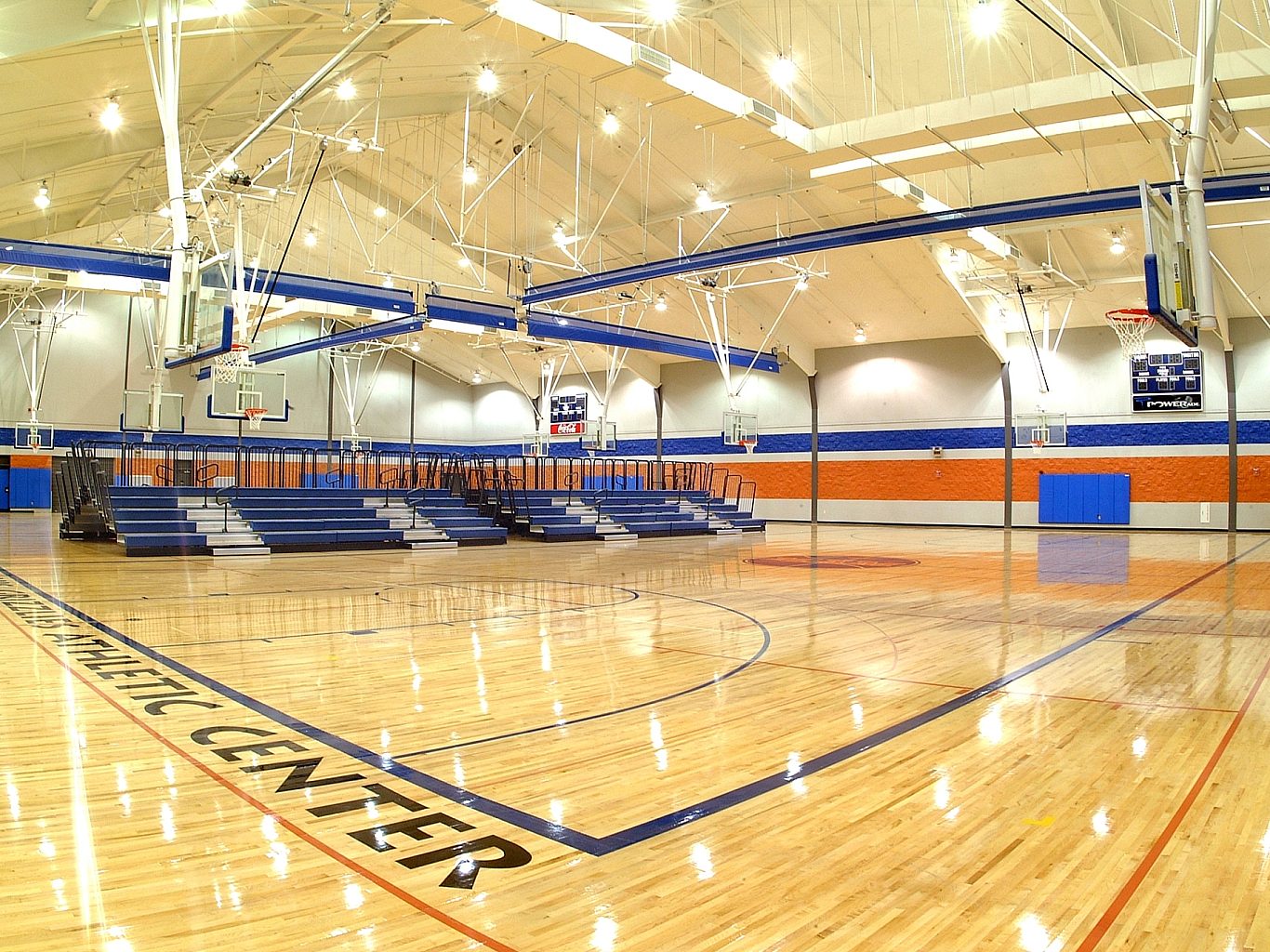 A basketball gym with dark blue bleachers along the far wall. Basketball goals hang from the ceiling, and Grizzlies Athletic Center is written across the floor.