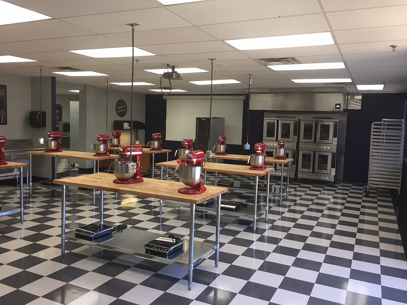 Wooden tables in the middle of a college culinary classroom that have two KitchenAid mixers on them each.