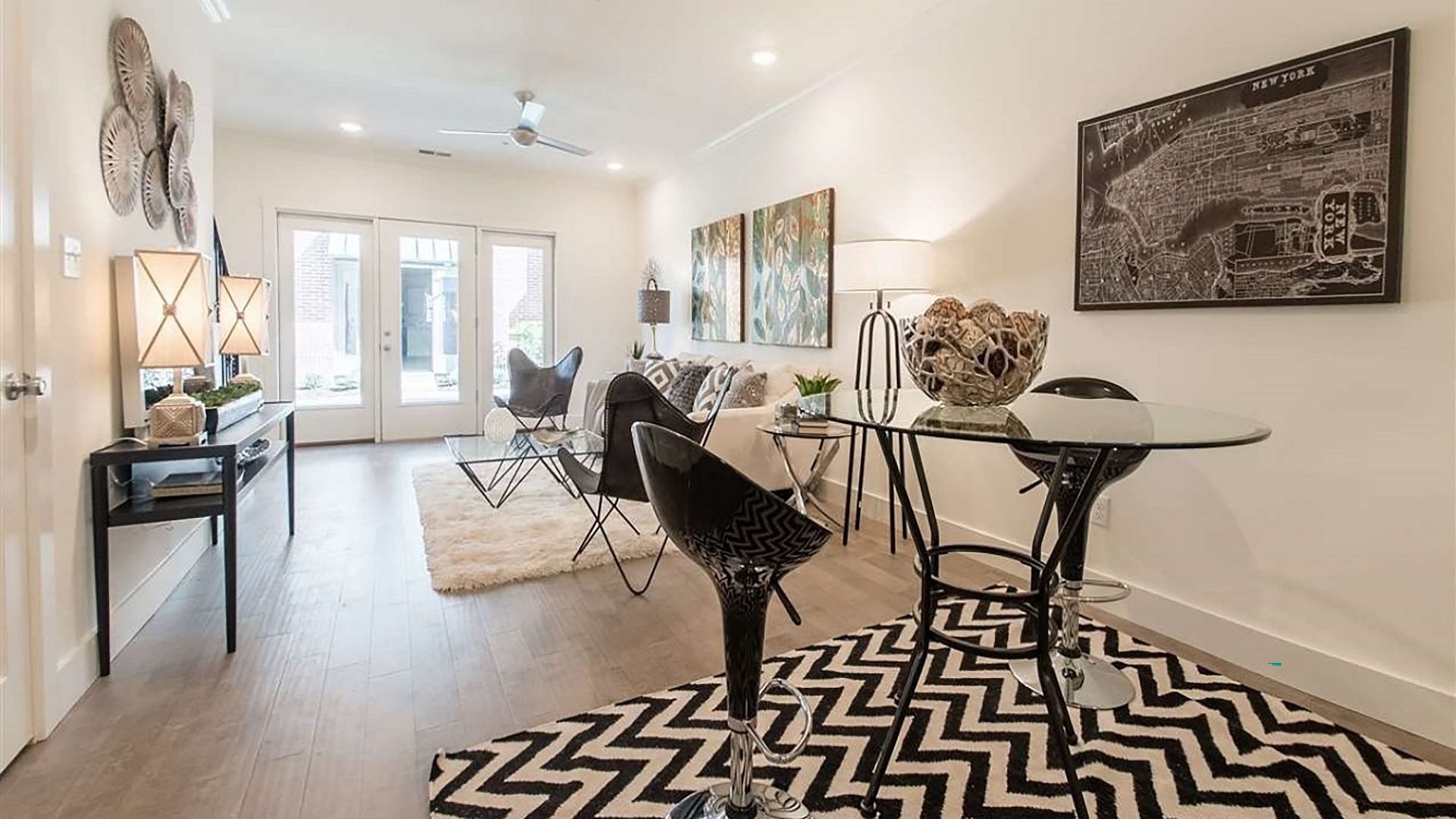 The living room of an apartment with modern furniture. A tall table with two chairs sits to the right, and artwork lines the walls.