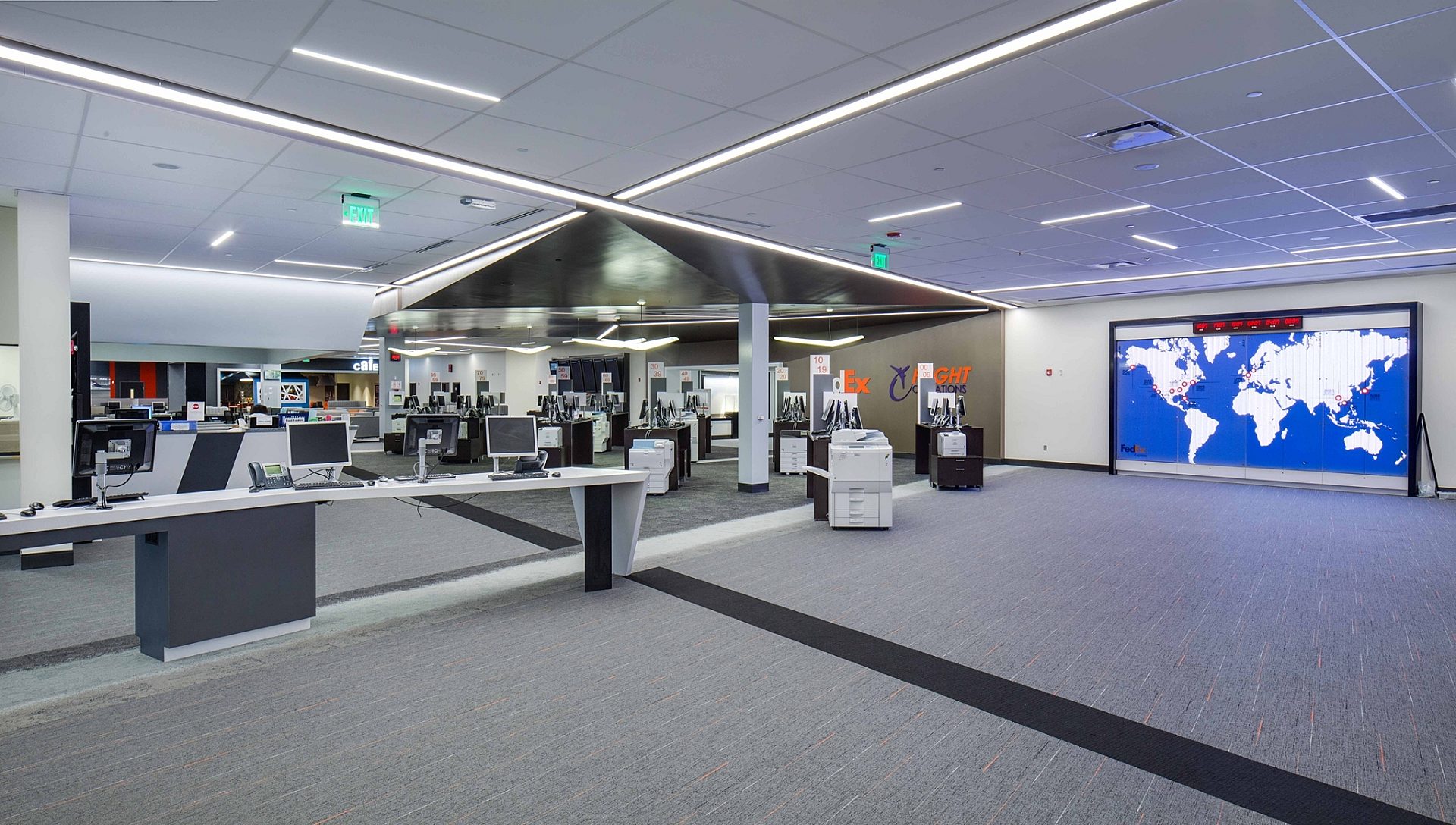 An office space with computers for use on a desk and printing stations on the right. A world map is on one wall.