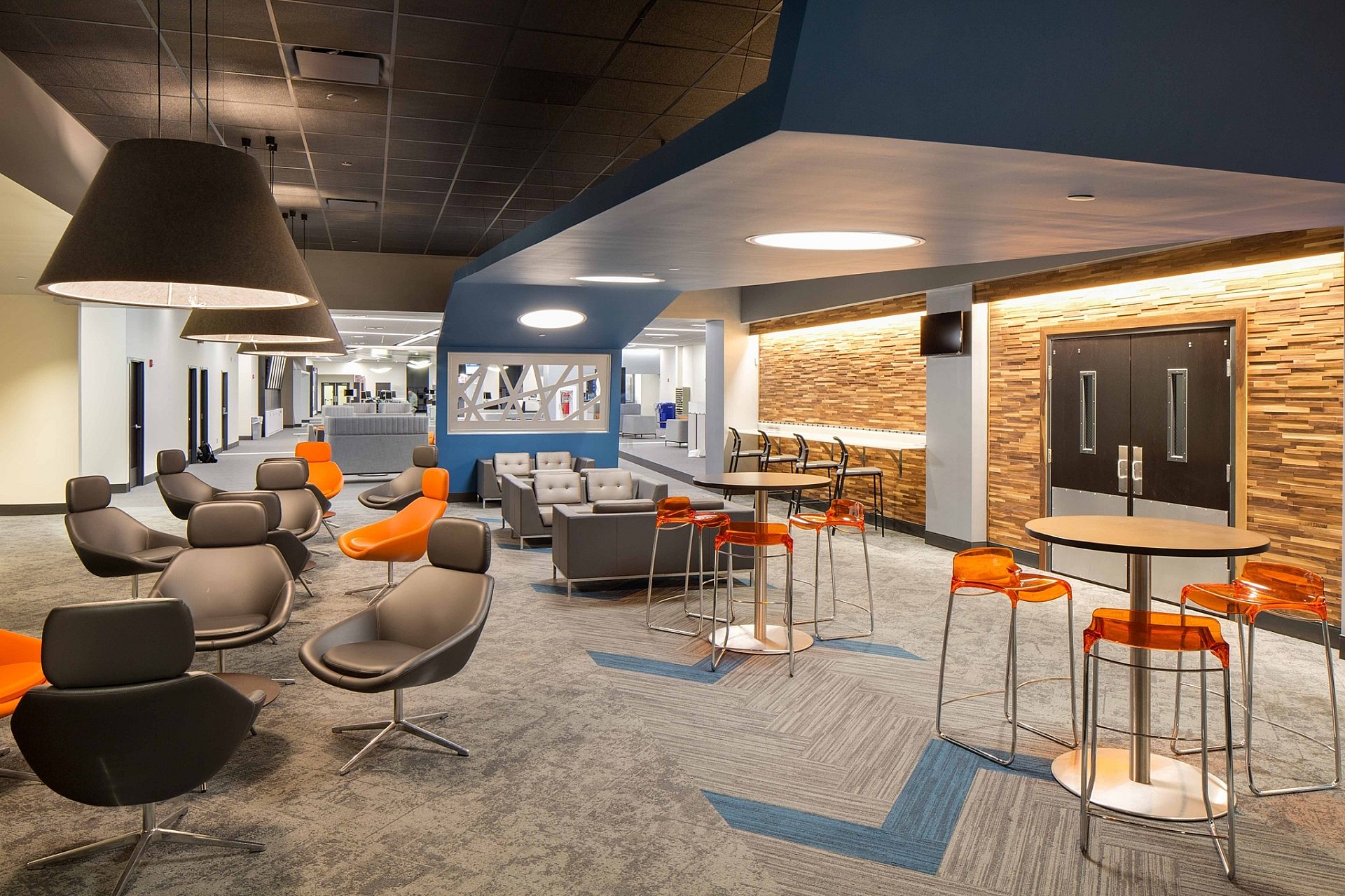 A common space in an office building with high top tables and orange bar stools mixed with black leather chairs. Wood paneling lines one wall.