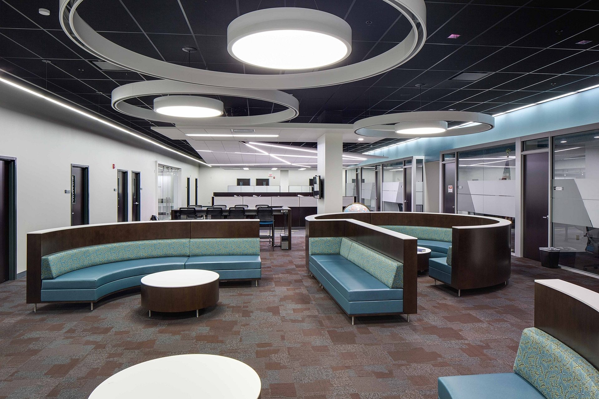 The common space of an office building with couches and tables in the middle. Round light fixtures hang from the ceiling.