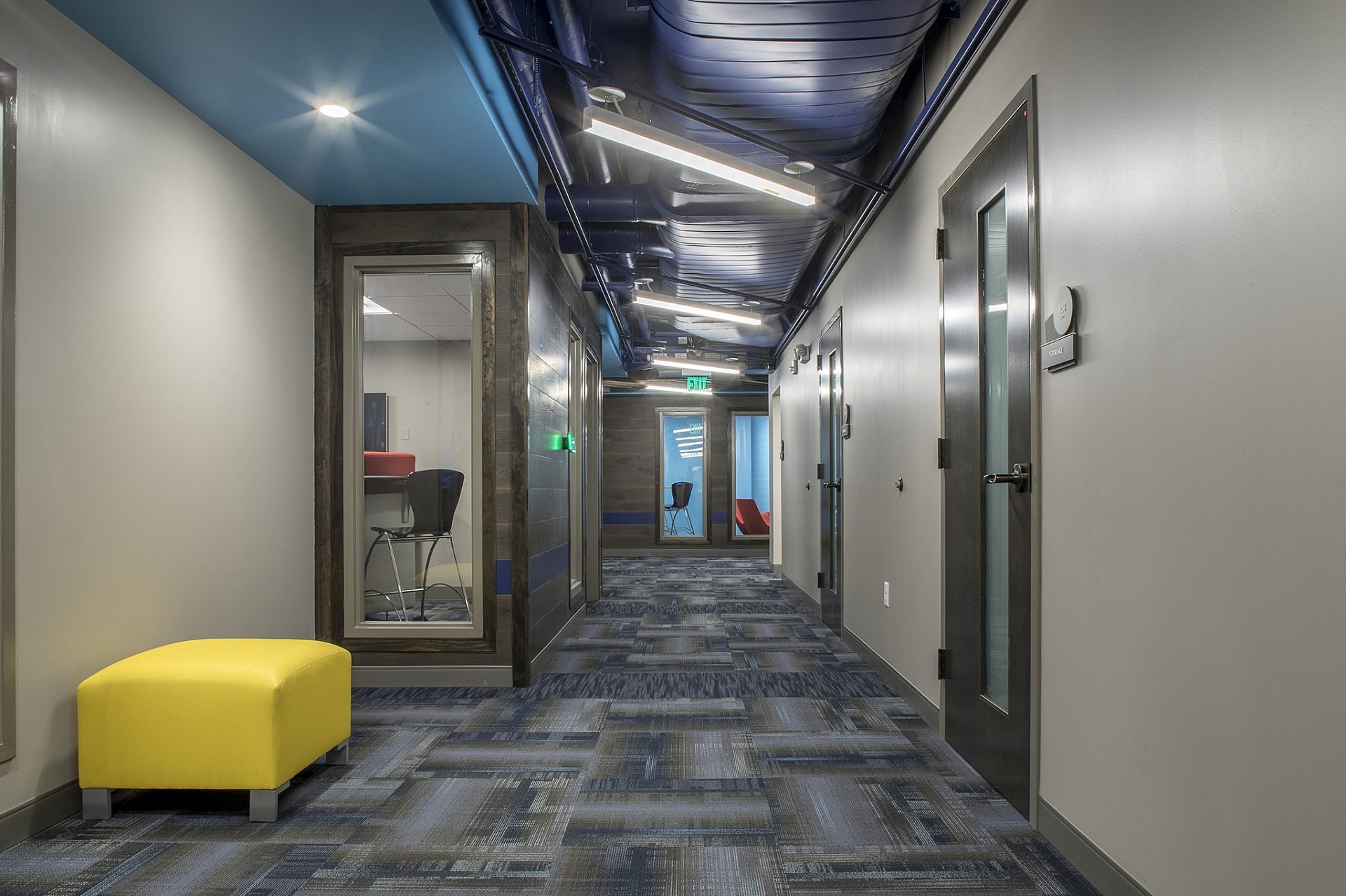 A hallway of an office building with a yellow ottoman on the left wall and offices seen through glass windows.