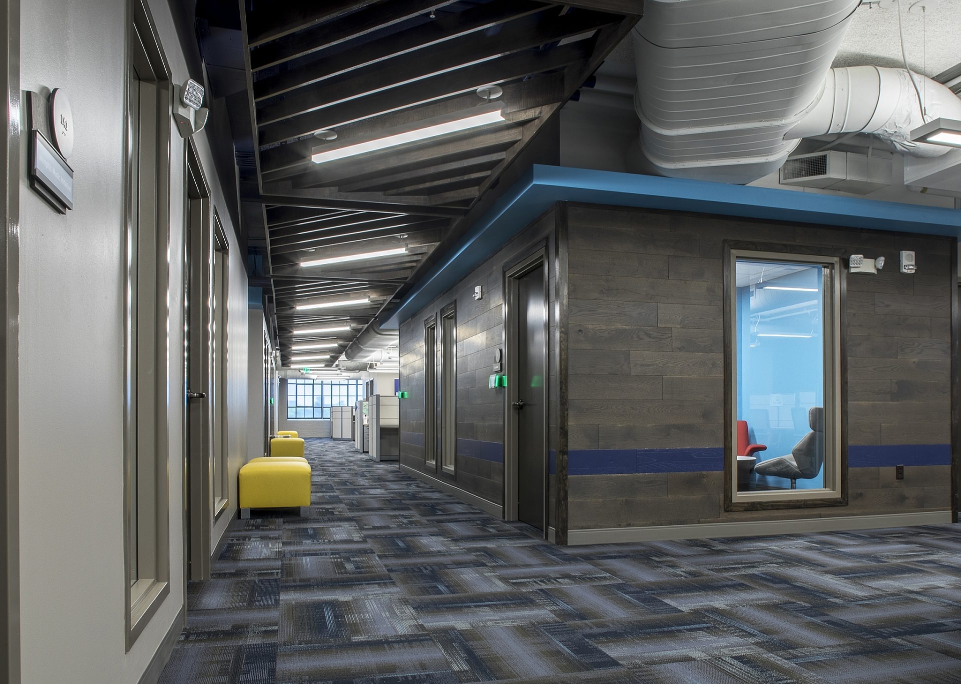 A hallway in an office building with yellow ottomans along the wall and office rooms on the right.