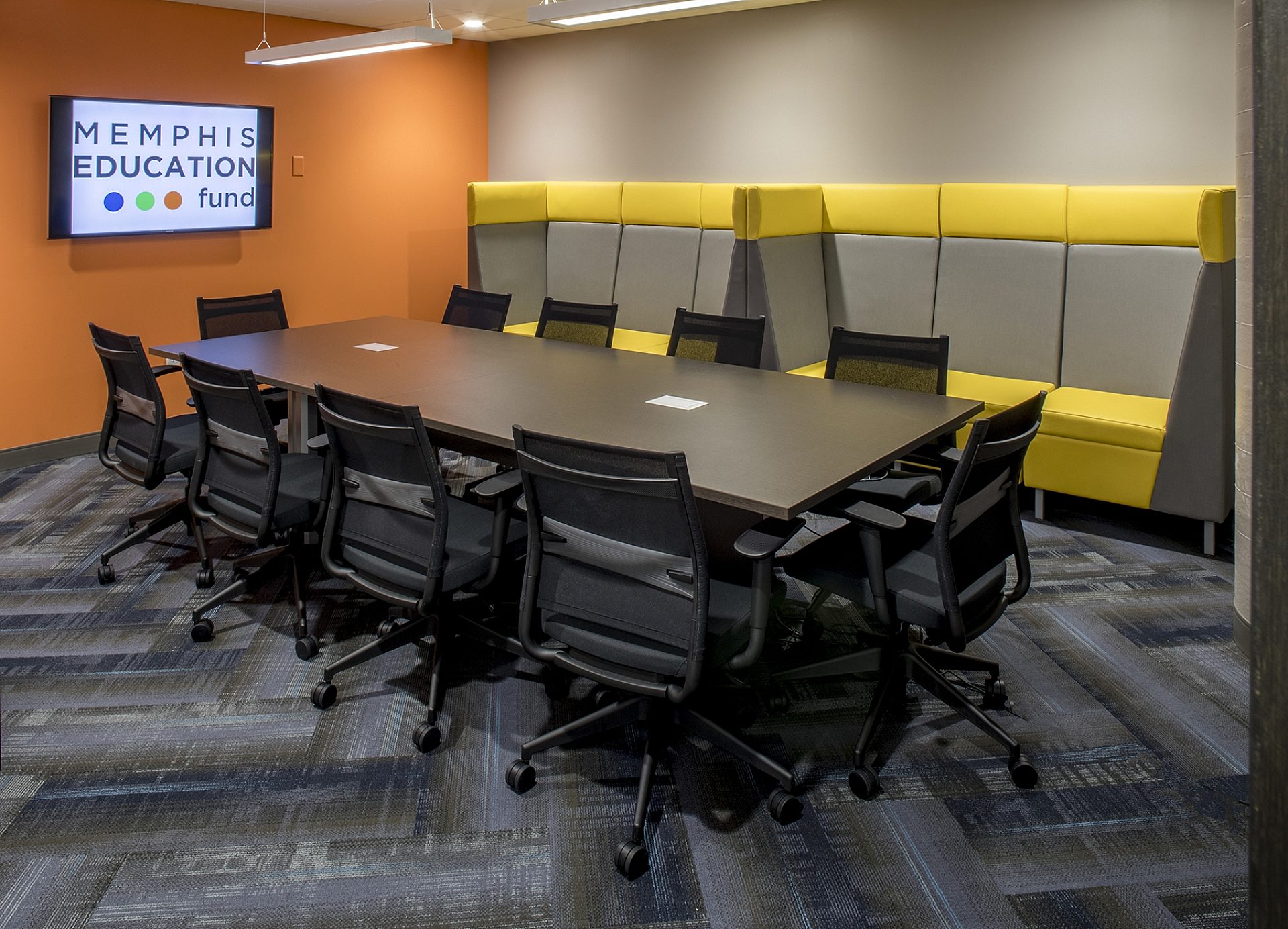A conference room with a rectangular table in the middle and chairs surrounding it. Seating lines the back wall.