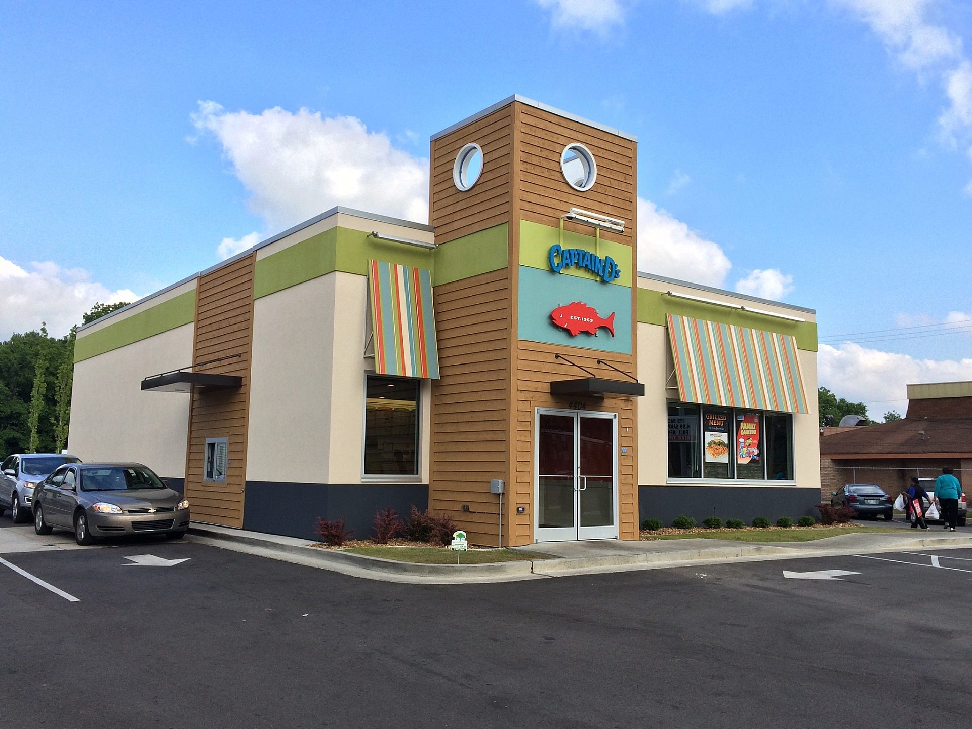 The exterior of a Captain D's restaurant. A red fish is above the entrance, and striped awnings are over the windows. A car is in the drive through.