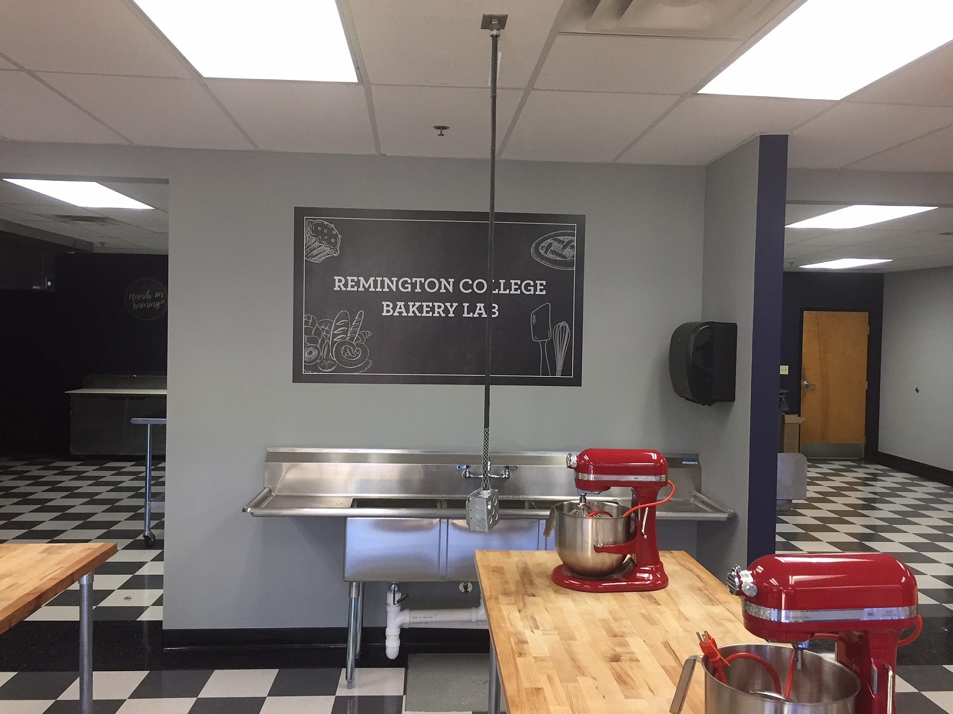 An industrial sink on the back wall of a college culinary classroom. A wooden table has two mixers on it.