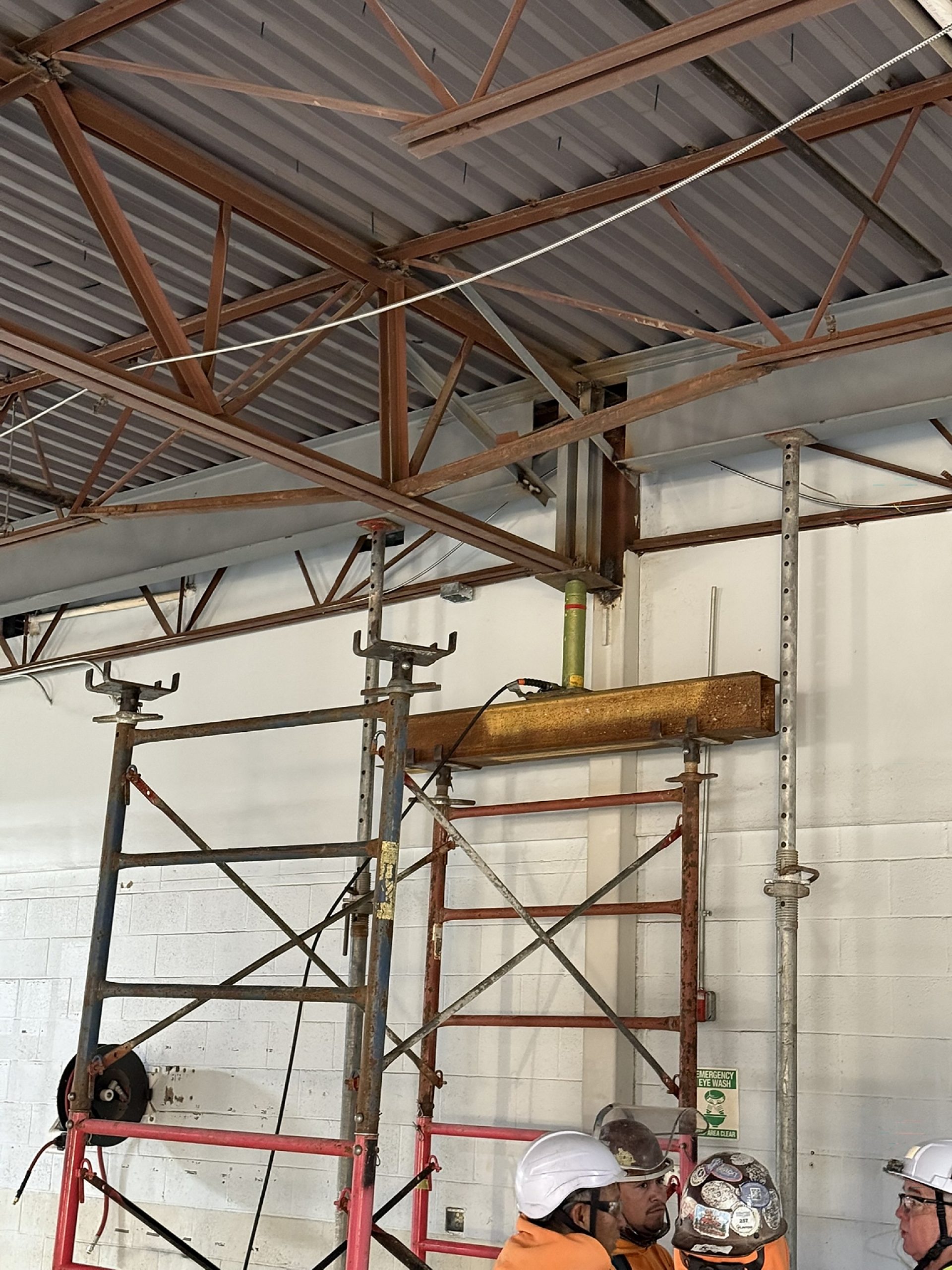 Interior construction scene showing steel roof trusses and beams overhead with construction workers in the foreground