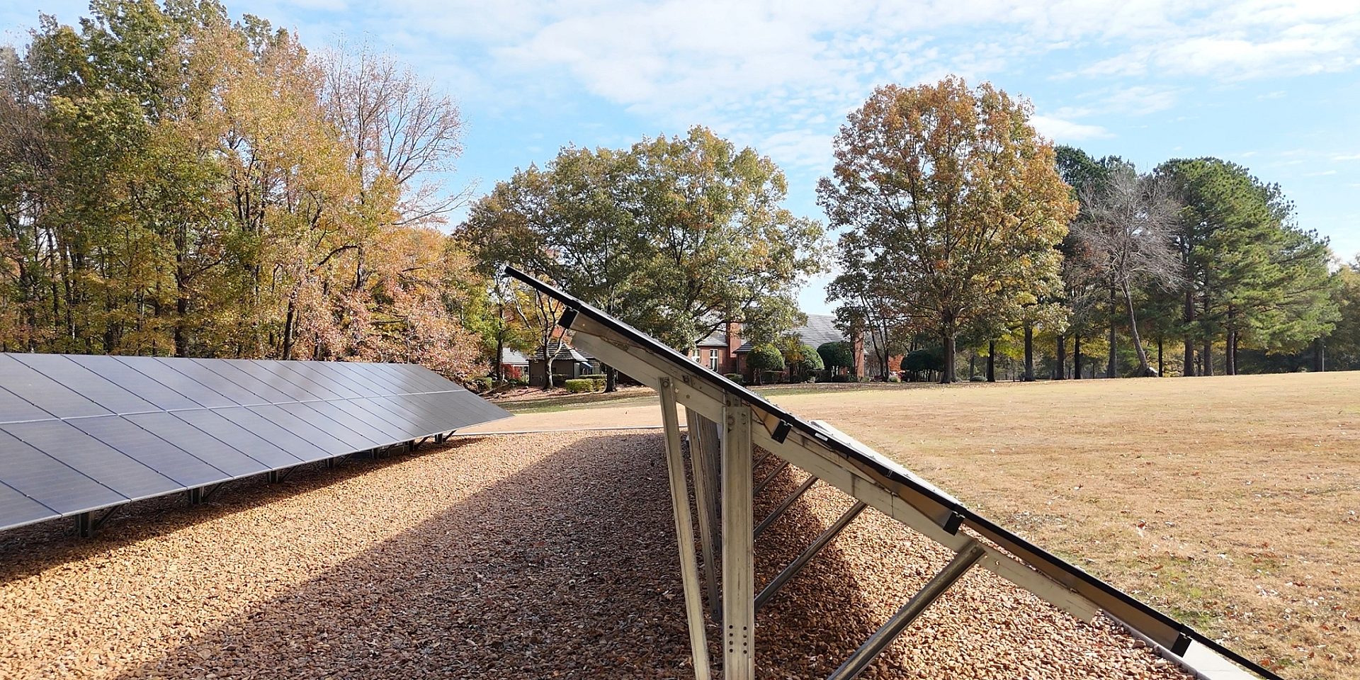 A side view of solar panels on a gravel pad with trees in the background.