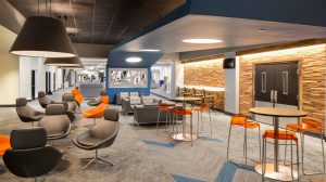 A common space in an office building with high top tables and orange bar stools mixed with black leather chairs. Wood paneling lines one wall.