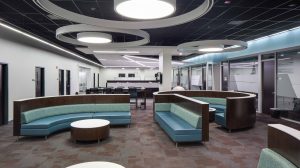 The common space of an office building with couches and tables in the middle. Round light fixtures hang from the ceiling.