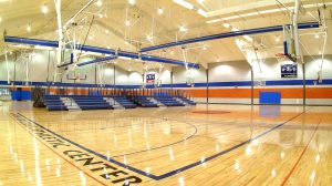 A basketball gym with dark blue bleachers along the far wall. Basketball goals hang from the ceiling, and Grizzlies Athletic Center is written across the floor.