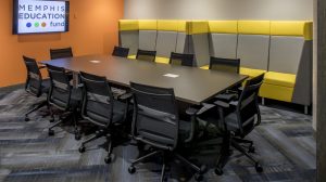 A conference room with a rectangular table in the middle and chairs surrounding it. Seating lines the back wall.
