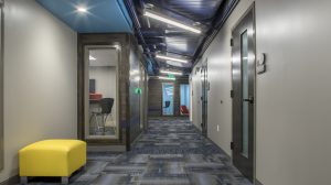 A hallway of an office building with a yellow ottoman on the left wall and offices seen through glass windows.