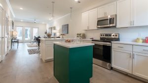 A view of a kitchen leading into the living room. An island is in the middle, and the oven is behind it in the middle of the built in cabinets.