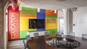 A conference room with a round table in the center and a colorful mural with a TV on the back wall.