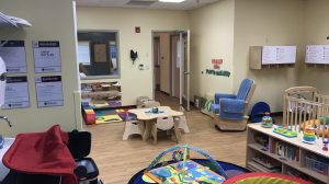 A classroom for toddlers with baby toys in the play area and a crib and rocking chair in the background.